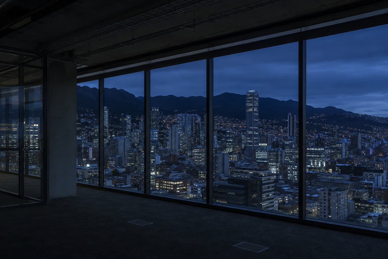 Planta de oficinas vacía tras un ventanal de piso a techo, con la ciudad encendida al anochecer y la cordillera al fondo.